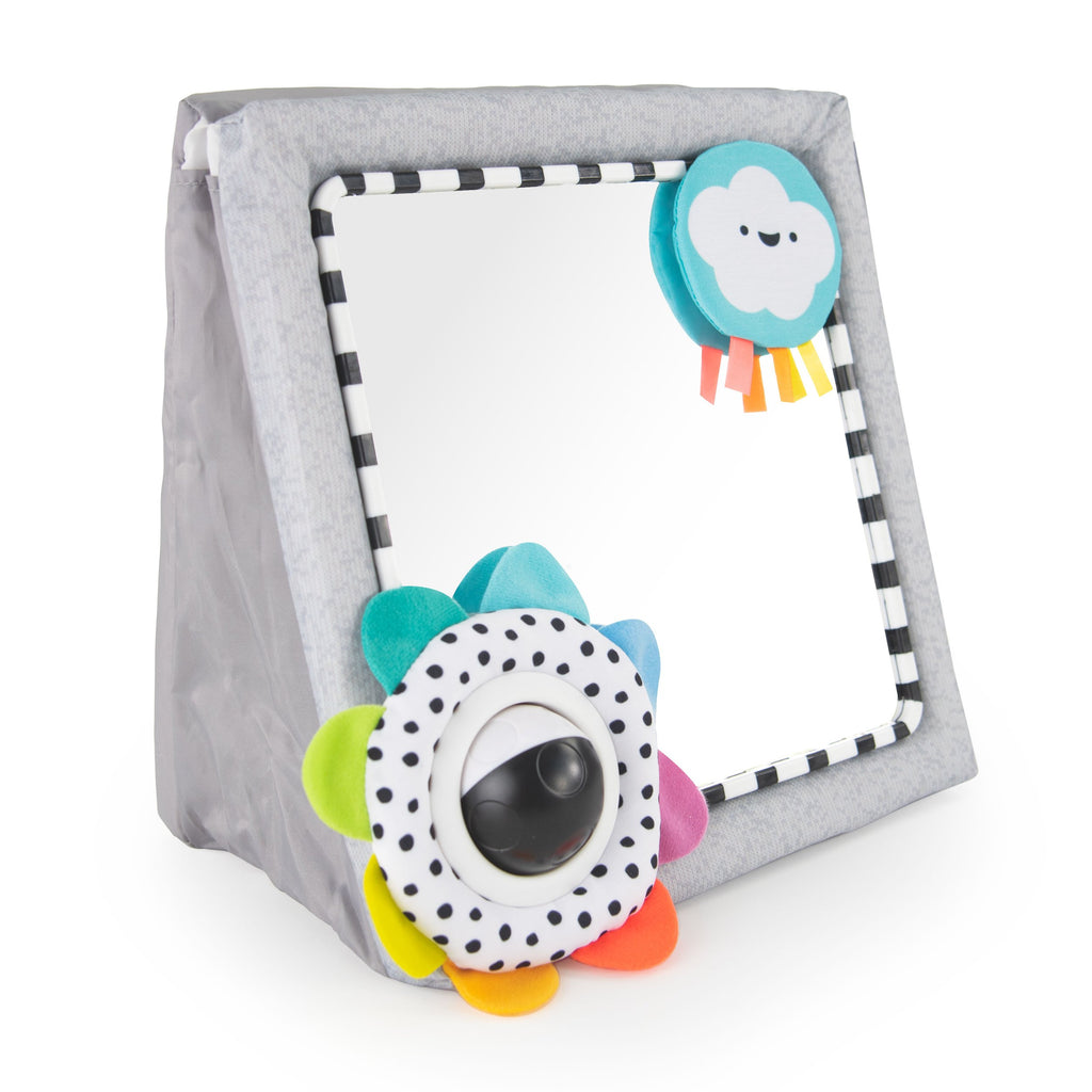 a wedge/triangle shaped floor mirror that can stand and lay flat for tummy time. the mirror has a black and white striped border. in the top right corner is a happy themed blue and white cloud with three ribbons attached at the bottom. in the bottom left corner is a black and white polka dot fabric attachment with a hard black and white spinning ball. there are rainbow ordered fabric petals around the polka dot attachment. 