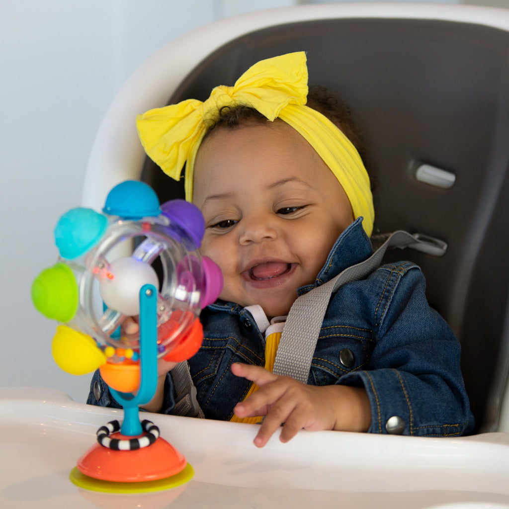 Baby playing with spinning wheel tray toy with multicolored bumps, attached to a suction base on a high chair.