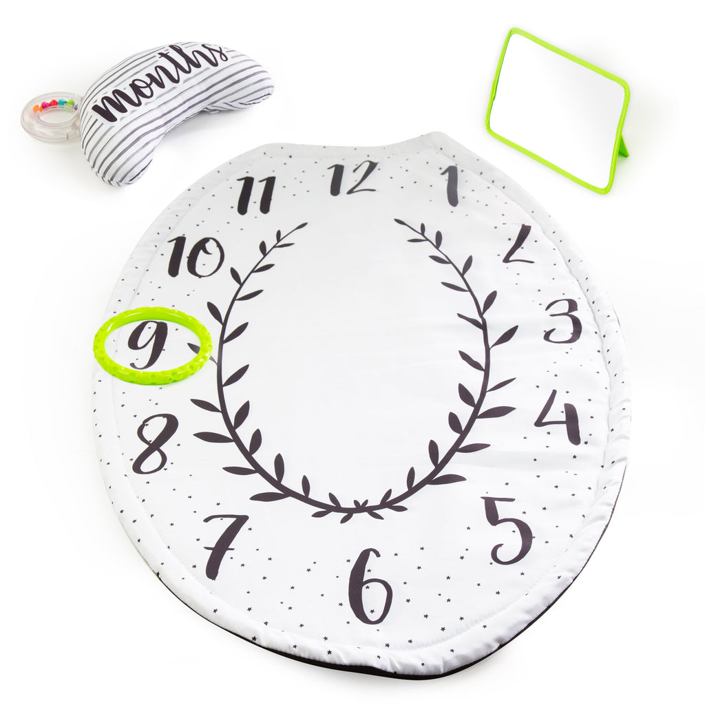 a white oval shaped tummy time play mat that has small star patterns around the border. the mat is numbered in a circle 1-12 with leaf pattern in the middle. there are three other pieces to the mat including mirror with a green border, a green ring to place on the numbers, and an arch shaped bolster with a striped pattern (months written across the top) with a clear ring filled with colored beads. 