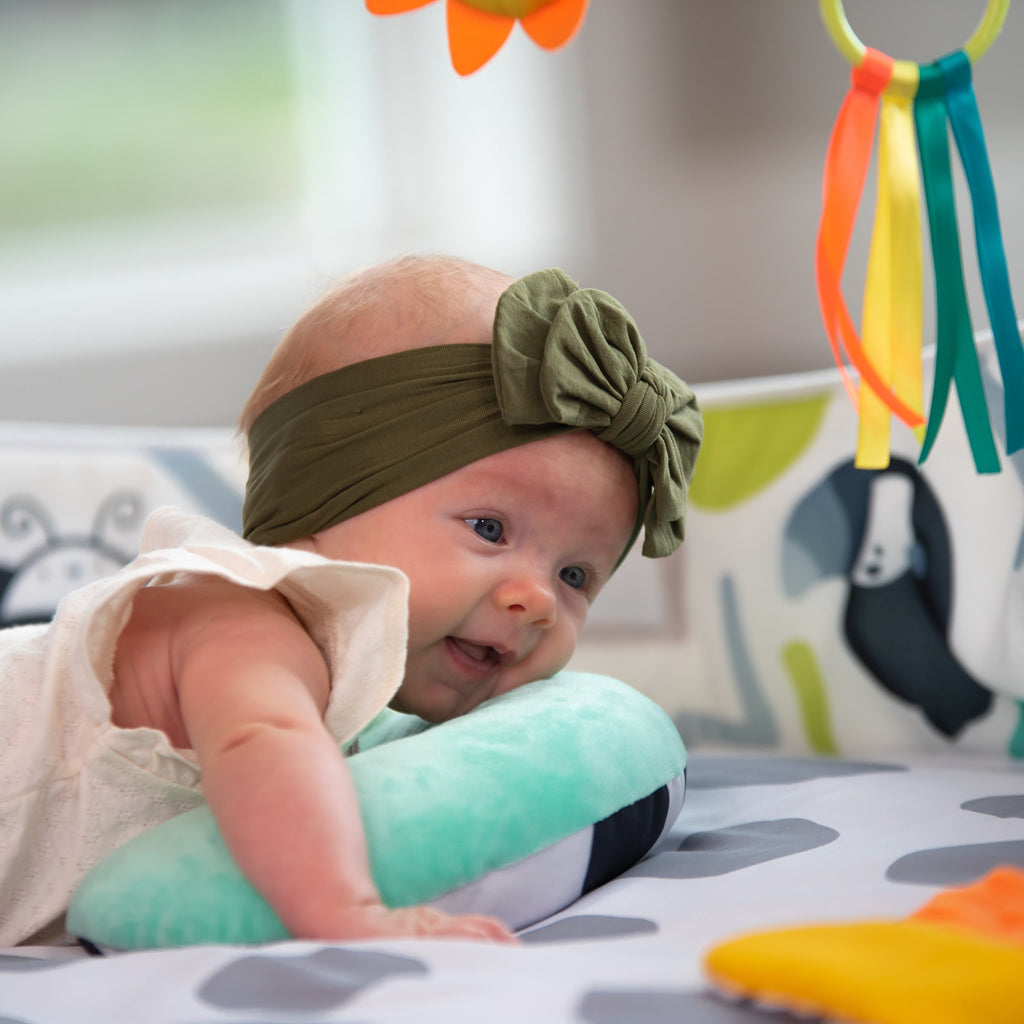 small baby with green bow on head laying on the bolster of the playmat to help with tummy time.