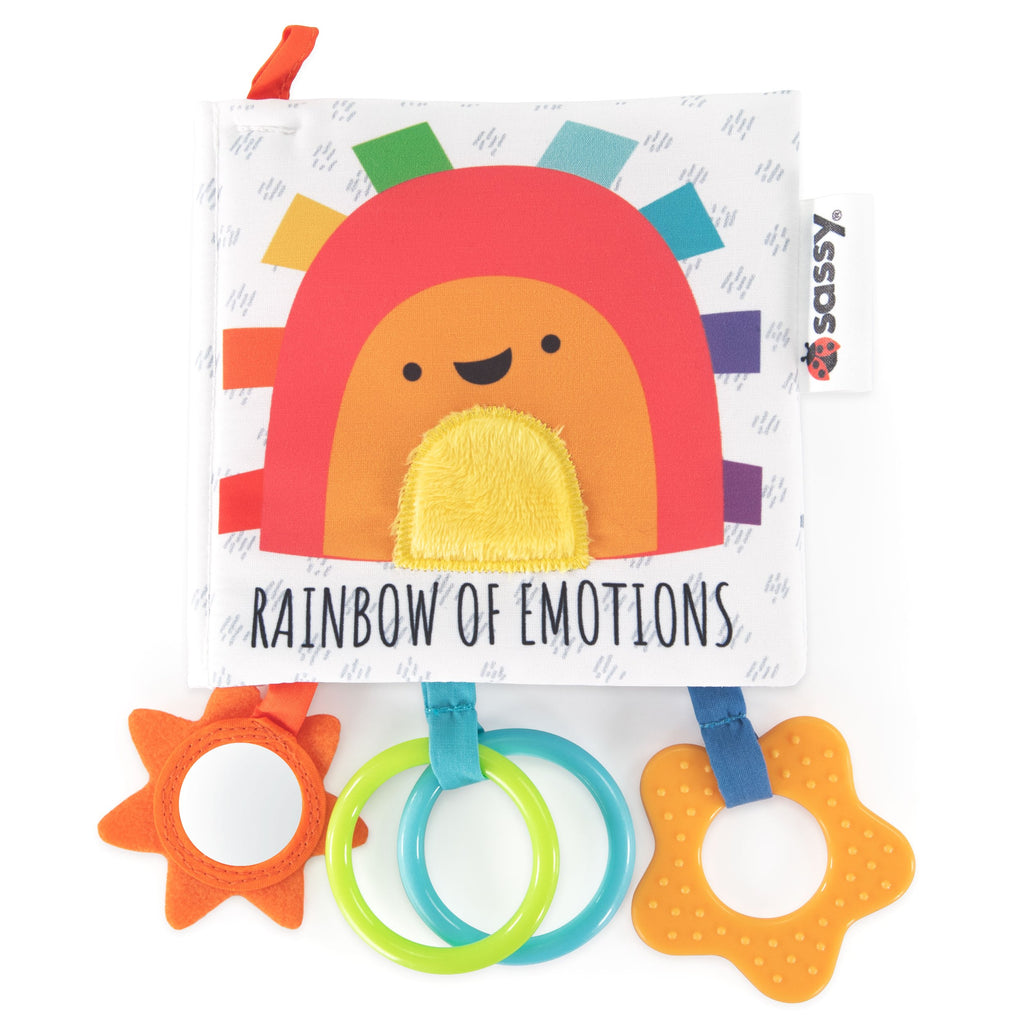 a soft square white book with crinkly light gray patterns and a rainbow character with ribbons on the cover, titled "rainbow of emotions." each page illustrates different emotions and characters in rainbow order (e.g., angry red mushroom, curious orange, careful yellow lemon, calm green pear, sad blue cloud, surprised purple grape, confused brown cookie). the book features three hanging activities: a sunshine mirror, a green/blue ring, and a textured yellow star.