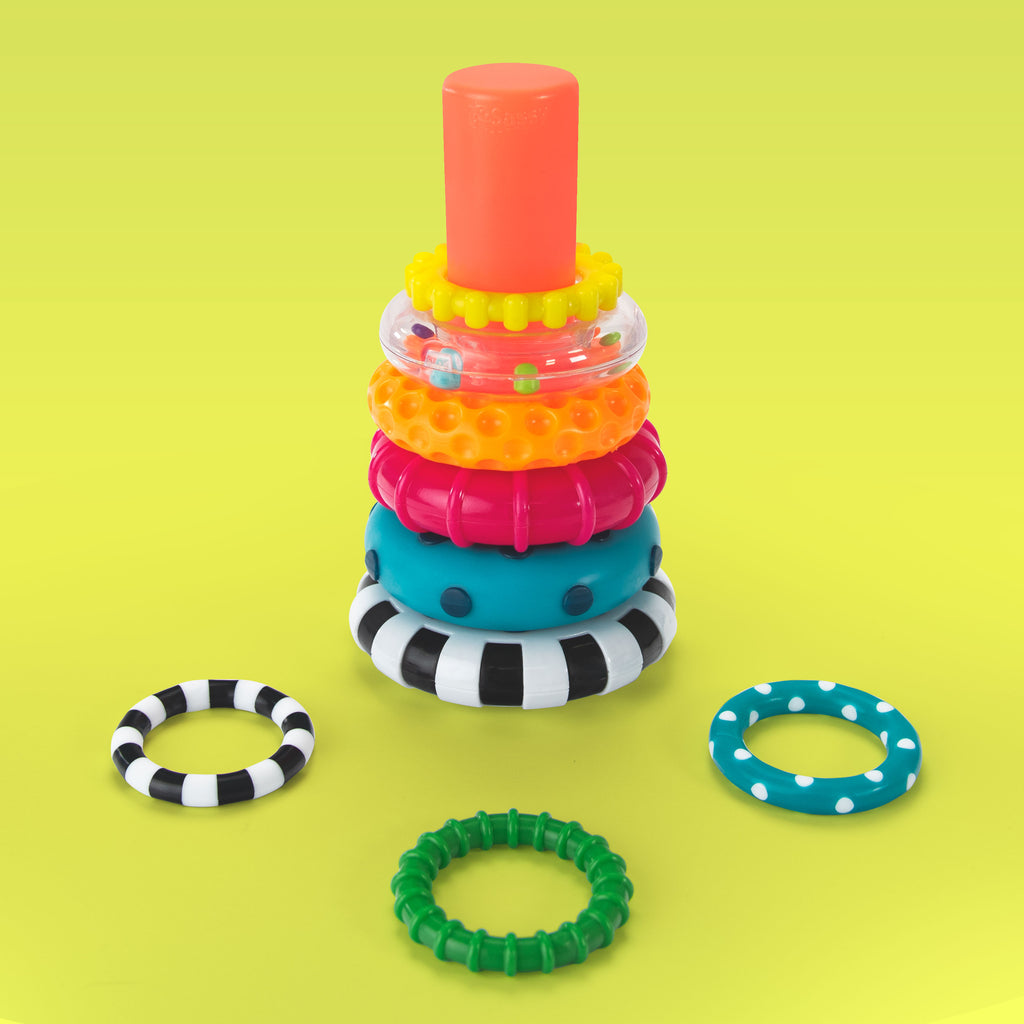 a black and white striped ring stacker base with a pink/red post featuring eight rings of various colors, textures, and sizes. the rings include a dark blue polka dot ring, a pink ring with textured stripes, an orange ring with inverted bumps, a clear ring filled with colored beads, a smaller yellow textured ring, a smooth blue and white polka dot ring, a small black and white striped ring, and a dark green ring