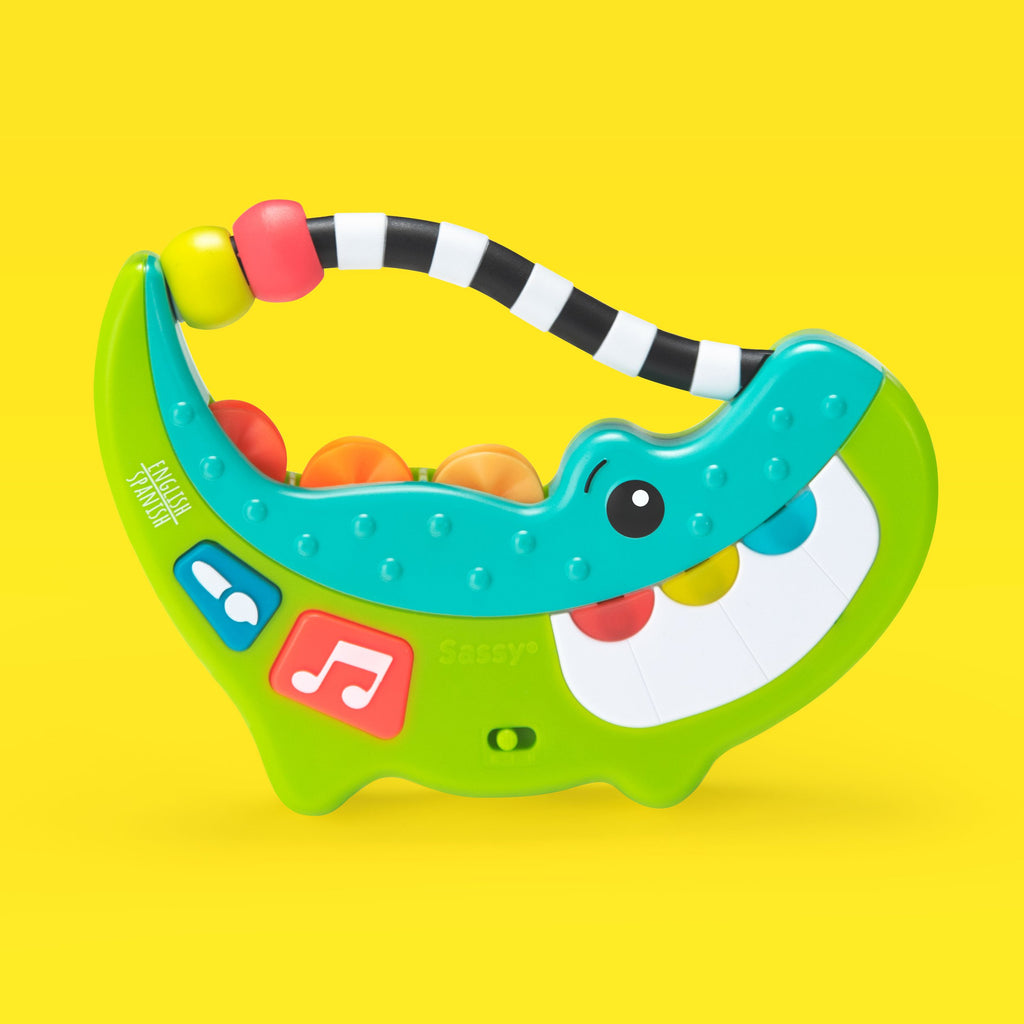 a green, blue, and white crocodile shaped musical toy that has a black and white piece attached at the top with two thick rings. the crocodile has two buttons on the left side with a musical note illustrated and a paint brush illustration that when you press it makes music and sounds. on top of the crocodile there are three multi-colored spinners. on the right side of the crocodile are teeth that you can press to make more sound. 