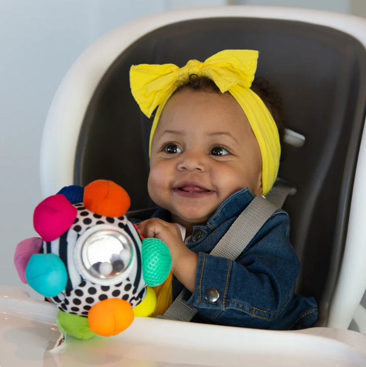 infant girl sitting in highchair, smiling, holding sassy bumpy ball toy