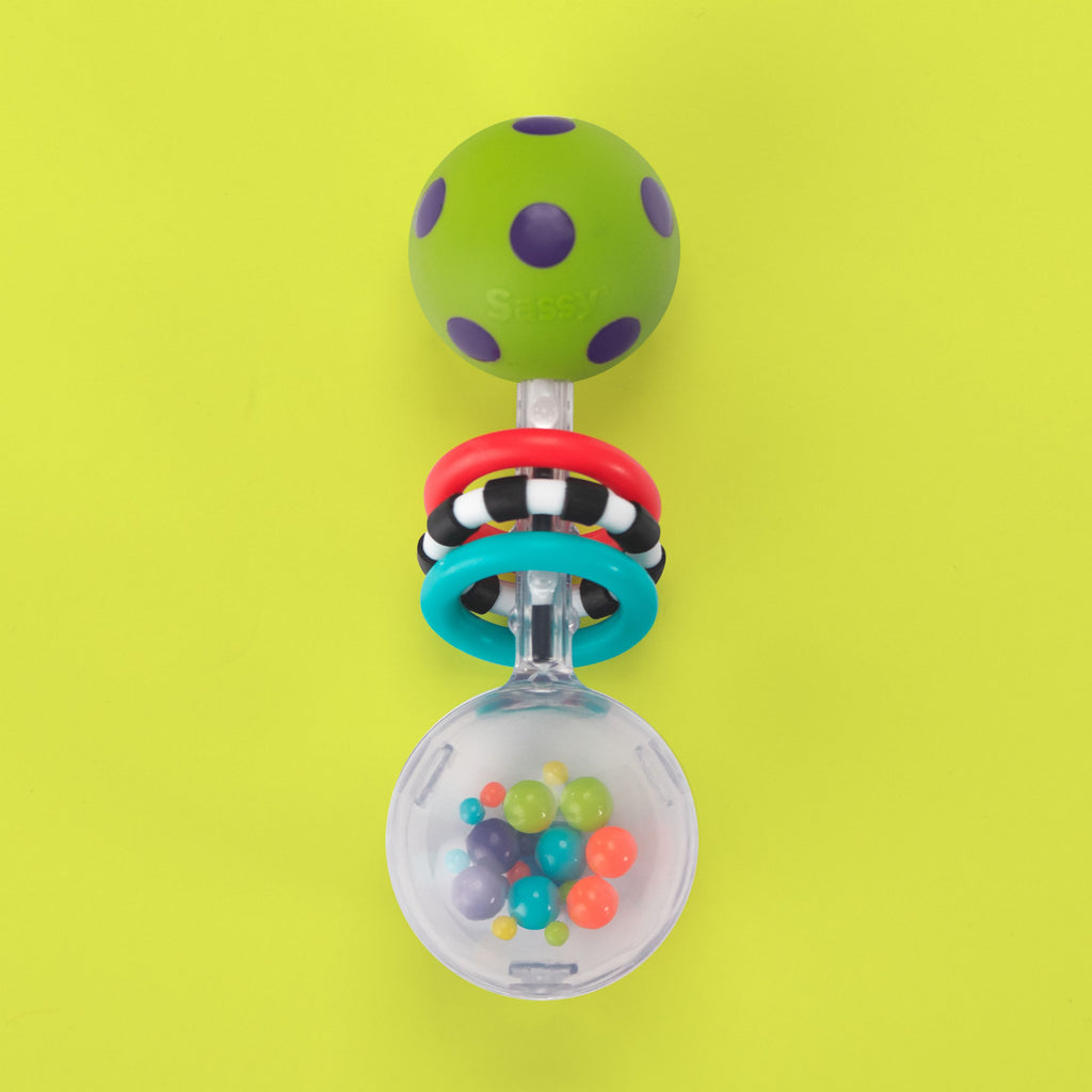 a stick rattle with a ball on both ends. one of the balls is green and purple polka dots with a smooth texture. the other ball is clear with multi-colored beads inside. there are three rings inbetween the balls. one ring is red with a smooth texture, the next ring is black and white striped with a ridged texture, and the last ring is blue with a smooth texture.