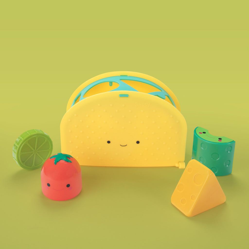 a taco shaped sorter with four different shaped slots. there are four different colored, textured shapes with faces. (red tomato, yellow cheese, green lime, green avocado slice).