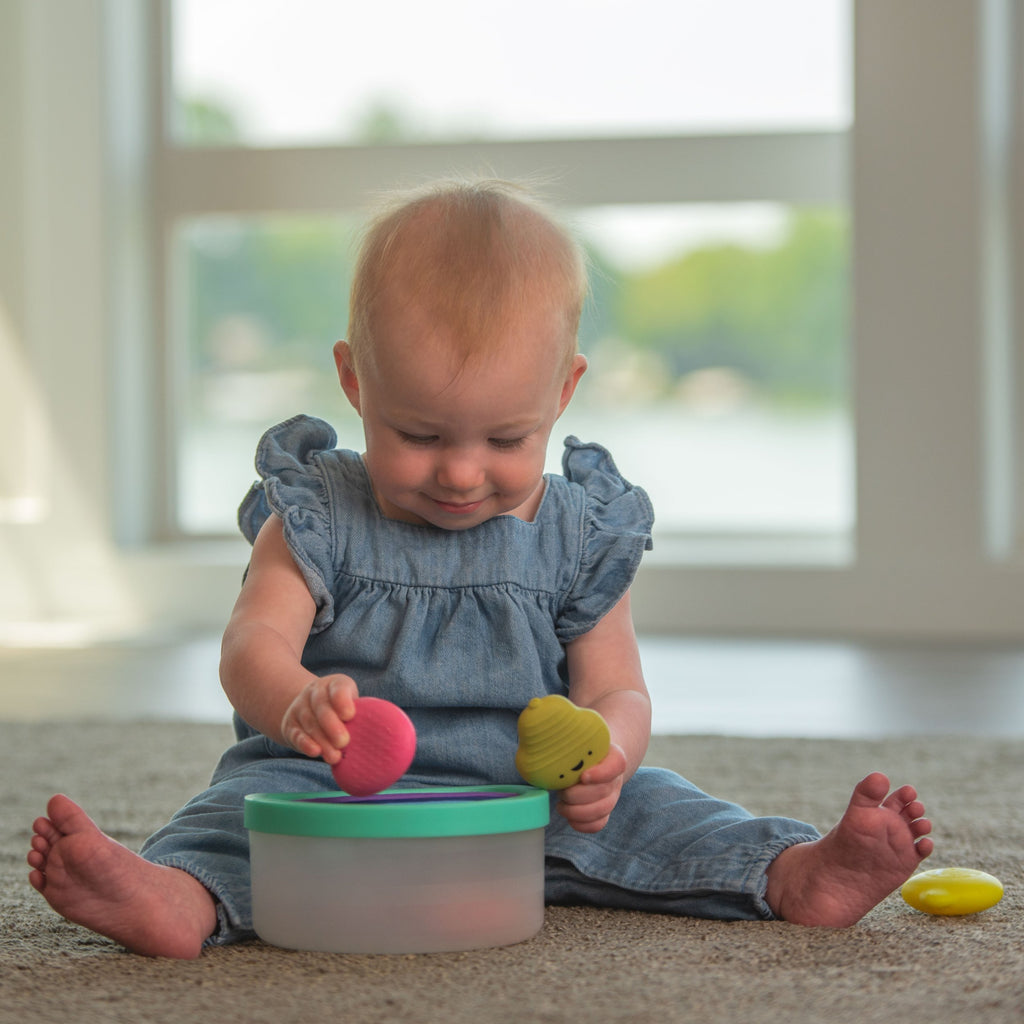 baby playing with fruit toys and placing them inside of a clear bin with a blue lid