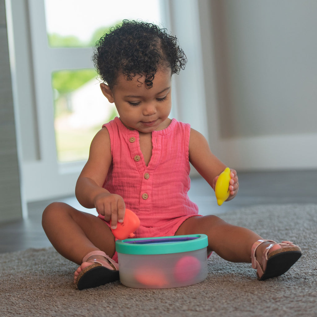 baby playing with fruit toys and placing them inside of a clear bin with a blue lid