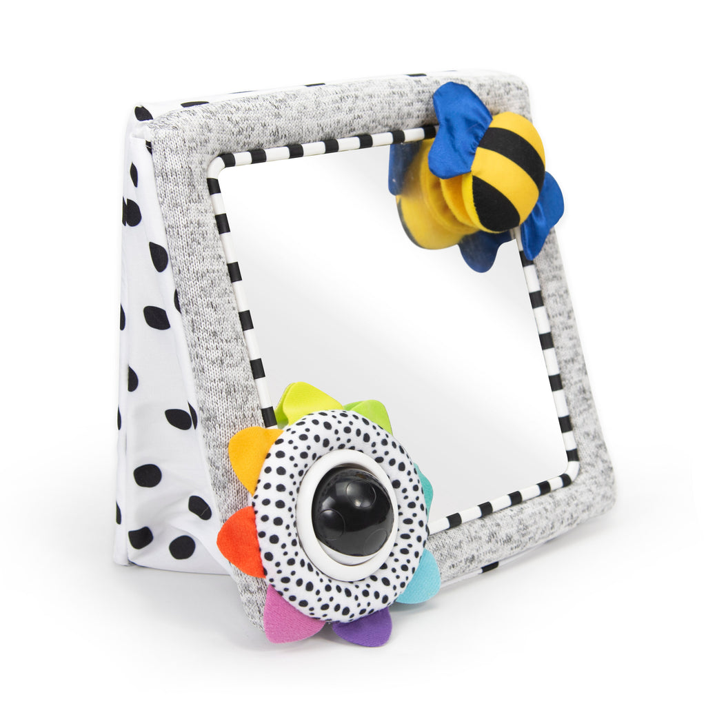 a triangle/wedge shaped tummy time mirror that can stand and lay flat. the back of the fabric is black and white polka dots. the mirror border is black and white striped. in the upper right corner is a black and yellow soft bee with blue wings. in the lower left corner is a circle black and white polka dot fabric piece with a black and white hard plastic ball inside. the border has rainbow ordered fabric petals. 