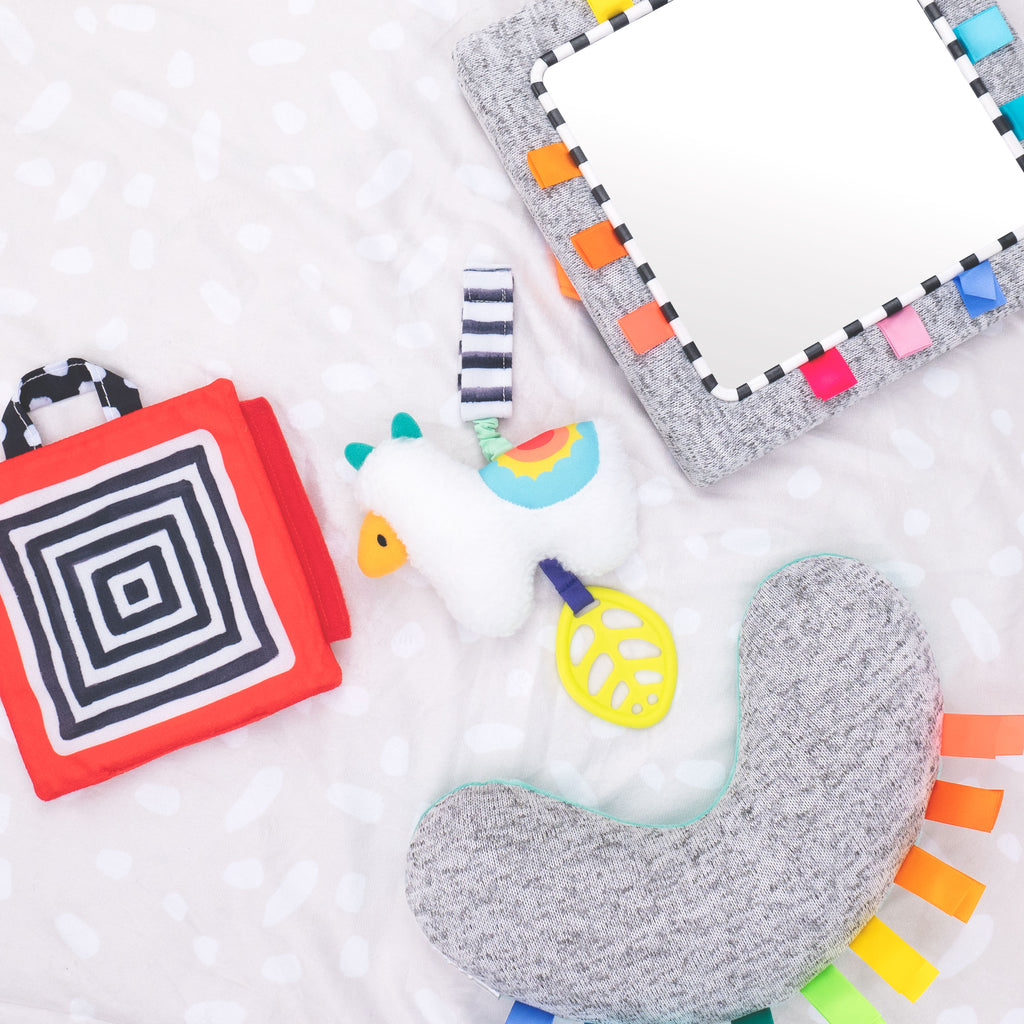 A grey bolster, a llama hanging toy, a black white and re sensory panel and a gray floor mirror all laying flat on a blanket.